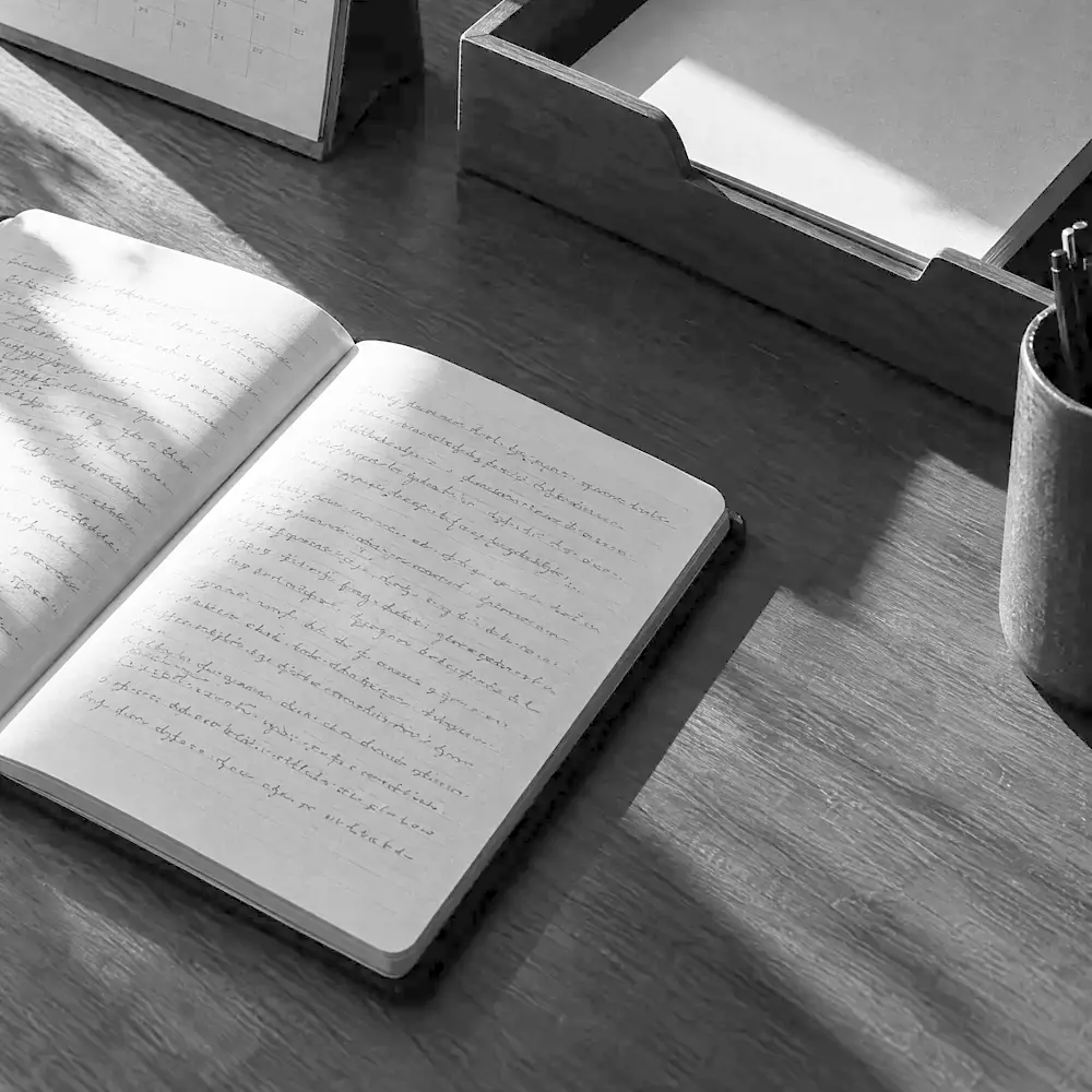 Aufgeschlagener Notizblock mit unleserlicher Handschrift, daneben Kalender, Papierablage und ein Becher mit Stiften auf einem Holztisch im harten Fensterlicht