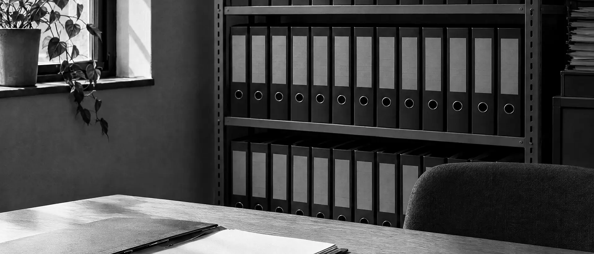 Regal mit Reihen von Aktenordnern in einem kleinen Büro, links ein Fenster, davor ein Schreibtisch mit aufgeschlagener Mappe